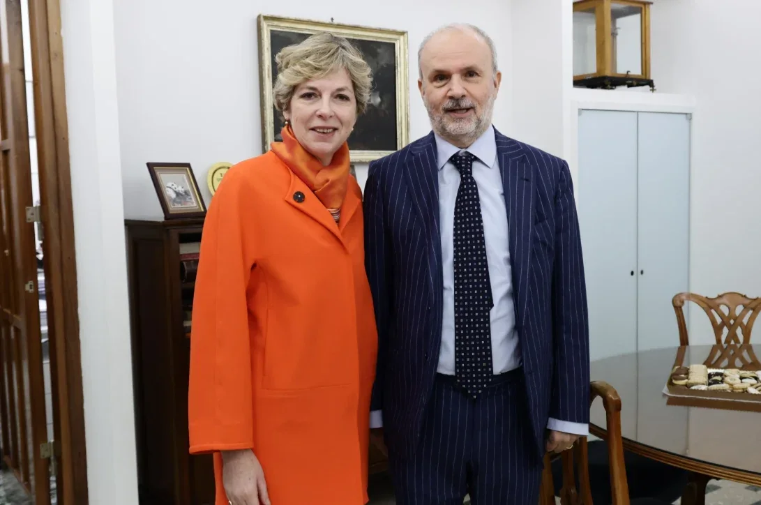 Orazio Schillaci in a pinstriped suit standing beside a woman in an orange coat in a formal office with period furniture and a framed portrait