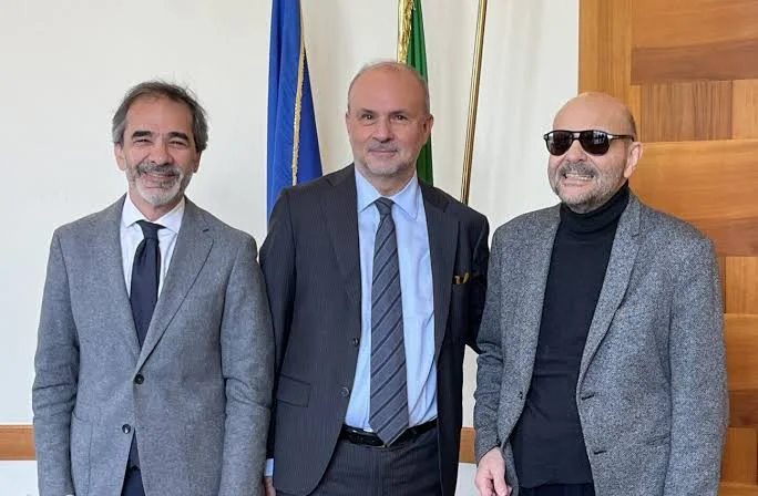 Orazio Schillaci standing between two men, one wearing dark glasses, in an office with the Italian and European flags