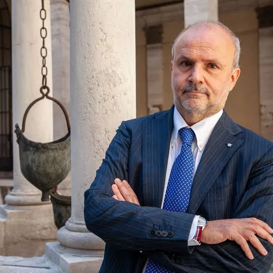Orazio Schillaci standing with arms folded in a stone cloister with classical columns and a hanging copper vessel
