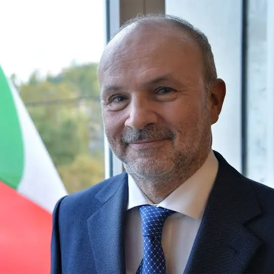 Orazio Schillaci smiling in a dark suit and patterned tie, with the Italian flag beside him