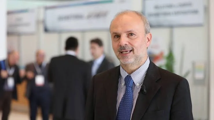 Orazio Schillaci smiling in a dark suit at a health congress, with exhibition stands and delegates in the background