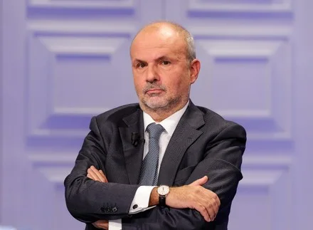 Orazio Schillaci standing with arms folded in a television studio with a lilac panelled backdrop