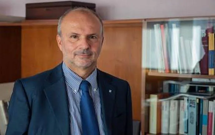 Orazio Schillaci in a navy suit and blue tie standing in front of wooden bookshelves filled with volumes