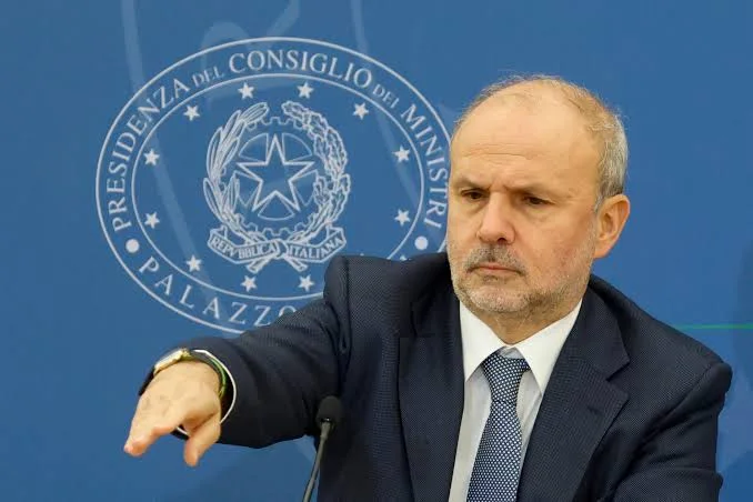 Orazio Schillaci gesturing at a press conference in front of the emblem of the Presidency of the Council of Ministers at Palazzo Chigi