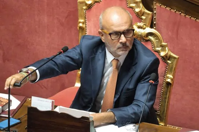 Orazio Schillaci seated in a red and gilded chair in a parliamentary chamber, adjusting a microphone